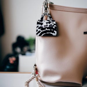 May include: A light brown tote bag with a black and white animal print charm attached. The charm has the text "NEW GENERATION" on it.