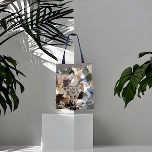 Può includere: Una borsa tote grigia con un design patchwork con vari colori e trame. La borsa ha manici blu scuro e il testo "NEW EDITION" è visibile. La borsa è esposta su un cubo bianco.