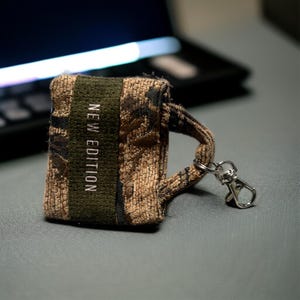 Könnte beinhalten: Ein kleiner Schlüsselanhänger aus grünem und braunem Tarnstoff mit einem silbernen Karabinerhaken. Der Stoff trägt den Aufdruck "NEW EDITION".