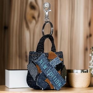 Könnte beinhalten: Ein kleiner Schlüsselanhänger in Form einer Jeans-Tragetasche mit schwarzem Band und silbernem Karabiner. Die Tasche ist aus verschiedenen Blautönen von Jeans gefertigt und hat einen braunen Leder-Patch mit dem Text "NEW EDITION" in Blau.