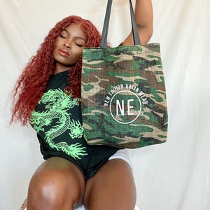 May include: A green and brown camouflage tote bag with black straps. The bag has a white circle with the text "NEW EDITION URBAN WEAR NE" on it.