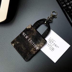 May include: A small black and gold tote bag keychain with a black strap and a silver clip. The bag has the words "NEW EDITION" printed on it in white.