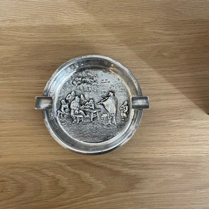 May include: Silver ashtray with a raised design of people gathered around a musician playing a fiddle. The ashtray has a slightly rounded shape and a lip around the edge.