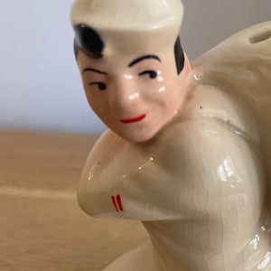 Op de afbeelding: Een keramiek beeldje van een matroos met een witte hoed en een wit shirt. Het beeldje zit in een hurkende positie met een gebarsten glazuur.