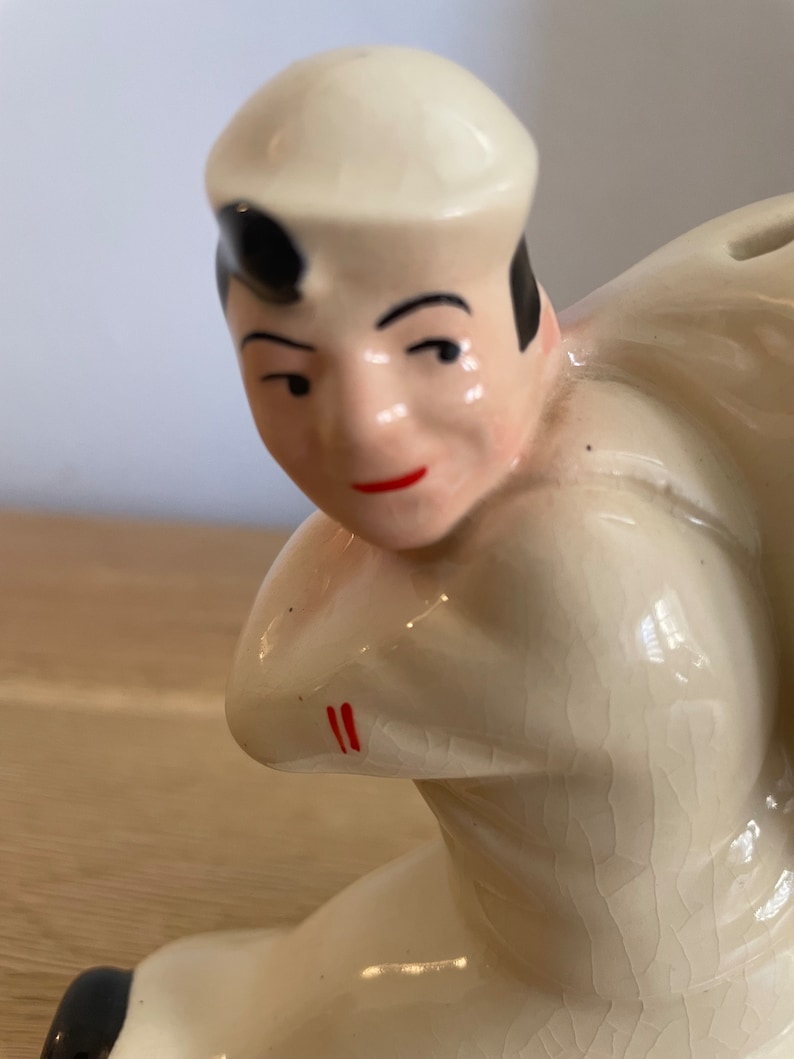 Op de afbeelding: Een keramiek beeldje van een matroos met een witte hoed en een wit shirt. Het beeldje zit in een hurkende positie met een gebarsten glazuur.