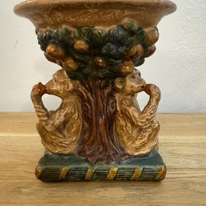 Peut inclure: Un pot décoratif en céramique de couleur marron et beige. Le pot présente un motif d'arbre avec des singes de chaque côté. L'arbre est chargé de fruits, et la base est de forme rectangulaire avec une bordure verte et dorée.