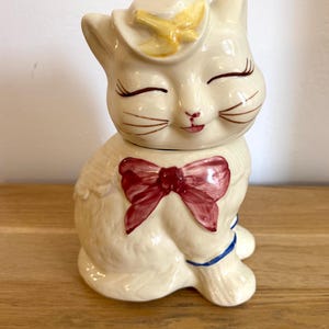 Pode incluir: Estatueta de gato em cerâmica creme com laço vermelho e detalhe amarelo no chapéu. O gato tem olhos fechados, boca vermelha pequena e detalhes azuis. Um item decorativo.