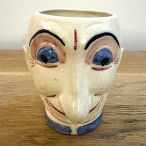Puede incluir: Una taza de cerámica vintage con forma de cara sonriente. La taza presenta ojos azules, detalles en rojo y un cuello azul. La cara está pintada sobre un fondo color crema, lo que le da un aspecto caprichoso y único.