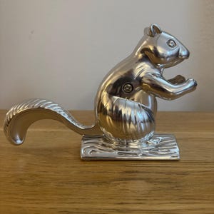 May include: A silver squirrel nutcracker. The nutcracker is shaped like a squirrel, with a curved tail and outstretched paws. It sits on a rectangular base with a wavy design. The nutcracker is made of metal.