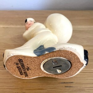 Op de afbeelding: Witte keramische spaarpot in de vorm van een persoon die een tas draagt. De spaarpot heeft een blauw en zwart ontwerp. De bodem van de spaarpot heeft een kurken basis met een metalen muntgleuf en de tekst "The Seamen's Bank for Savings".