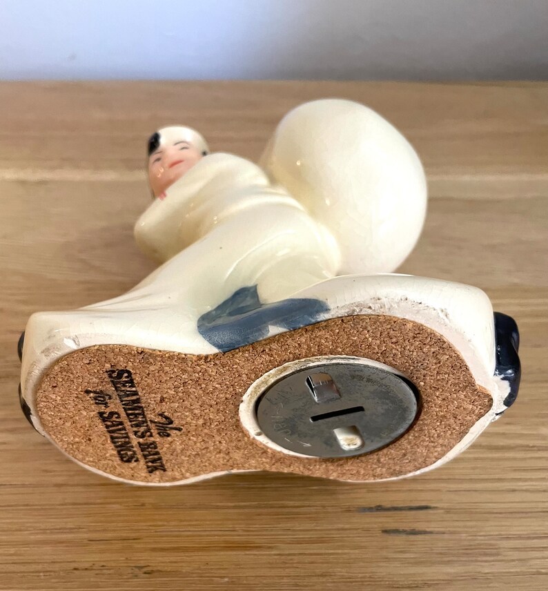 Op de afbeelding: Witte keramische spaarpot in de vorm van een persoon die een tas draagt. De spaarpot heeft een blauw en zwart ontwerp. De bodem van de spaarpot heeft een kurken basis met een metalen muntgleuf en de tekst "The Seamen's Bank for Savings".