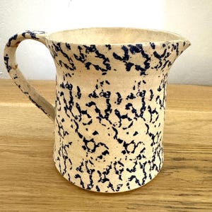 Può includere: Una brocca in ceramica con base color crema e un motivo blu scuro maculato o dipinto a spugna su tutto l'esterno. Il recipiente presenta un manico curvo sul lato sinistro e un piccolo beccuccio per versare sul bordo superiore destro.