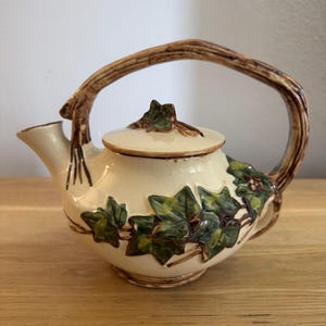 May include: A small, cream-colored teapot with a brown handle and a lid decorated with green ivy leaves.