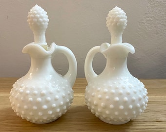 Puede incluir: Dos botellas de vidrio blanco con asas y tapones. Las botellas tienen forma bulbosa y están cubiertas de pequeñas protuberancias en relieve. Los tapones tienen forma de cono con una superficie texturizada. Los cruets se exhiben sobre una superficie de madera.