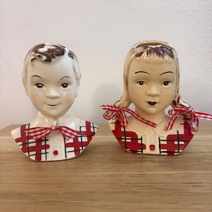 May include: Two vintage ceramic head vases, one depicting a boy and the other a girl. The boy has brown hair and a red plaid bow tie. The girl has pigtails tied with red plaid bows. Both have red plaid shirts.