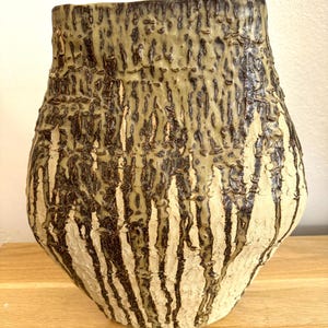 May include: A ceramic vase with a textured, bark-like surface. The vase is a light beige colour with brown and green glaze dripping down the sides.