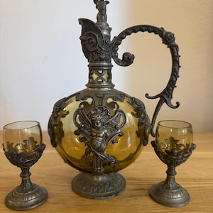 Puede incluir: Un juego de decantador de vidrio antiguo con dos vasos de chupito a juego. El decantador tiene un cuerpo de vidrio teñido de amarillo con adornos de metal, incluido un motivo de ángel. Los vasos de chupito tienen un diseño similar. El juego se exhibe sobre una superficie de madera.