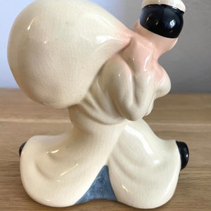 Op de afbeelding: Een keramiek beeldje van een persoon die een grote zak op zijn rug draagt. Het beeldje is geschilderd in tinten wit, roze en zwart. De persoon draagt een witte hoed en een wit shirt. De zak is wit en heeft een getextureerd oppervlak.
