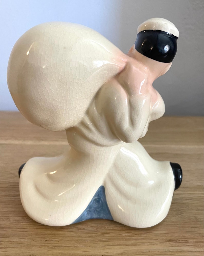 Op de afbeelding: Een keramiek beeldje van een persoon die een grote zak op zijn rug draagt. Het beeldje is geschilderd in tinten wit, roze en zwart. De persoon draagt een witte hoed en een wit shirt. De zak is wit en heeft een getextureerd oppervlak.