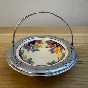 Puede incluir: Un pequeño cuenco de cerámica blanca con un diseño floral colorido se encuentra dentro de una fuente de servir plateada con asa. La fuente tiene un borde decorativo calado.