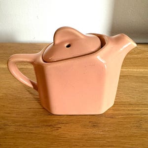May include: A pale pink ceramic teapot with a lid and a handle. The teapot has a square shape with rounded edges.