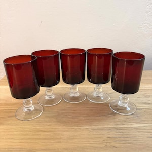 Puede incluir: Conjunto de cinco vasos de vidrio rojo con tallo. Los vasos son transparentes con un tinte rojo y tienen una base redonda.