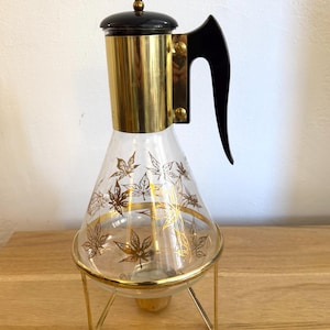 May include: Vintage glass coffee carafe with gold accents and a black handle. The carafe features a gold maple leaf design and sits on a gold metal stand. The top is black with a gold handle.
