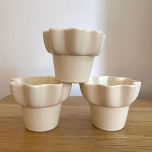 Pode incluir: Três vasos de flores de cerâmica creme com uma borda ondulada. Os vasos são empilhados em uma superfície de madeira. Os vasos têm um design simples e elegante, adequado para pequenas plantas ou suculentas.
