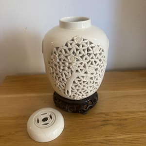 Könnte beinhalten: Cremefarbene Keramikvase mit baumartigem Ausschnittmuster, auf einem schwarzen Holzständer. Die Vase hat einen passenden Deckel mit kreisförmigem Muster. Das Design zeigt florale und geometrische Formen.