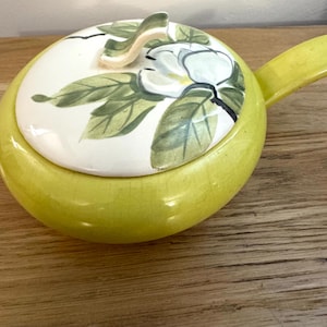 May include: A yellow ceramic serving dish with a lid. The lid is white with a floral design of white flowers and green leaves. The dish has a handle and is sitting on a wooden surface.