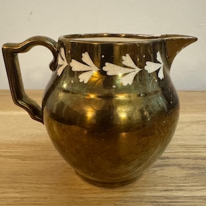 Könnte beinhalten: Eine goldfarbene Vintage-Kanne mit gebogenem Griff und Ausguss. Die Kanne hat ein weißes Blumenmuster am Rand. Die Kanne ist wahrscheinlich aus Keramik oder Porzellan und eignet sich zum Servieren von Getränken.