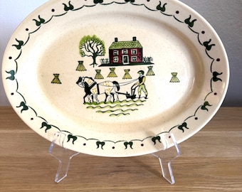 Puede incluir: Plato ovalado de cerámica con fondo crema y borde decorativo verde y rojo. El centro presenta una escena de granja con una casa roja, un árbol y un granjero arando con caballos. Una pieza encantadora para servir.