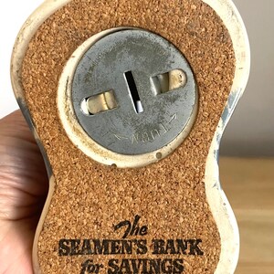 Op de afbeelding: Een wit keramiek spaarpotje in de vorm van een zeeman met een kurken bodem. Het spaarpotje heeft een metalen muntgleuf met de tekst "Turn" erop gedrukt. Op de bodem van het spaarpotje staat "The Seaman's Bank for Savings".
