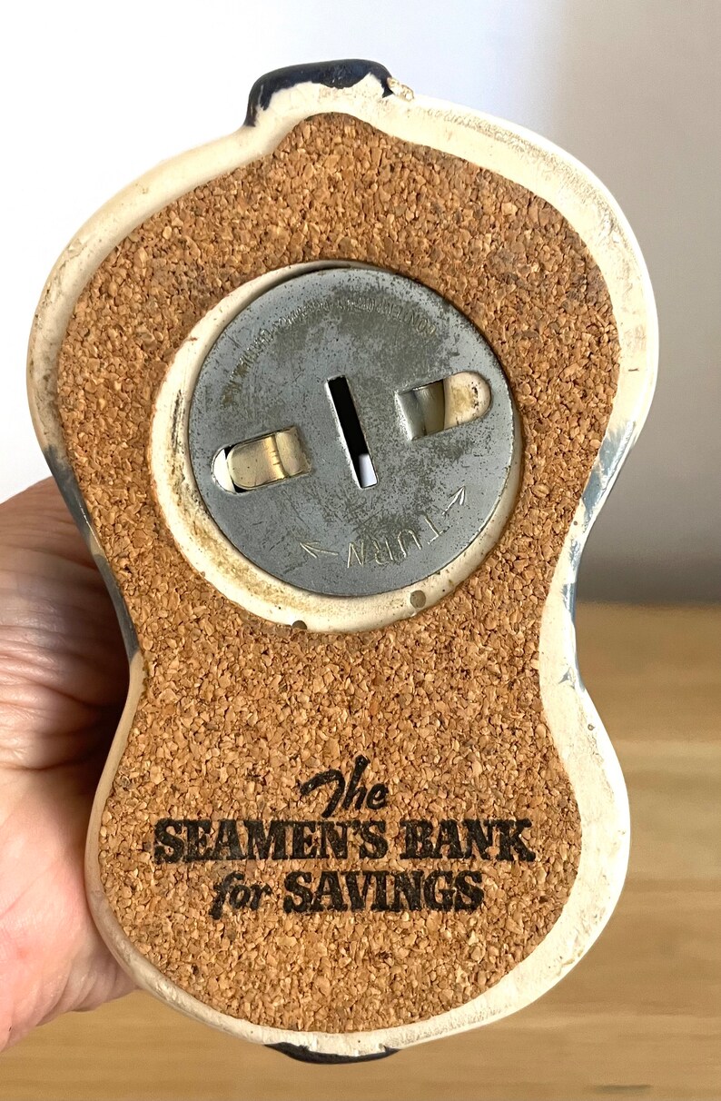 Op de afbeelding: Een wit keramiek spaarpotje in de vorm van een zeeman met een kurken bodem. Het spaarpotje heeft een metalen muntgleuf met de tekst "Turn" erop gedrukt. Op de bodem van het spaarpotje staat "The Seaman's Bank for Savings".