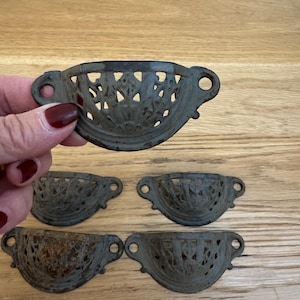 May include: Set of five antique-style drawer pulls. Each pull is a dark, weathered metal with a decorative, openwork design. The pulls have a curved shape with small holes and circular mounting points on each end. The pulls are on a wooden surface.