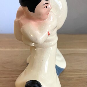 Op de afbeelding: Een keramiek beeldje van een matroos met een witte hoed, zwart haar en een wit uniform. De matroos houdt een groot wit voorwerp in zijn rechterhand. Het beeldje is geschilderd met rode accenten op het uniform.