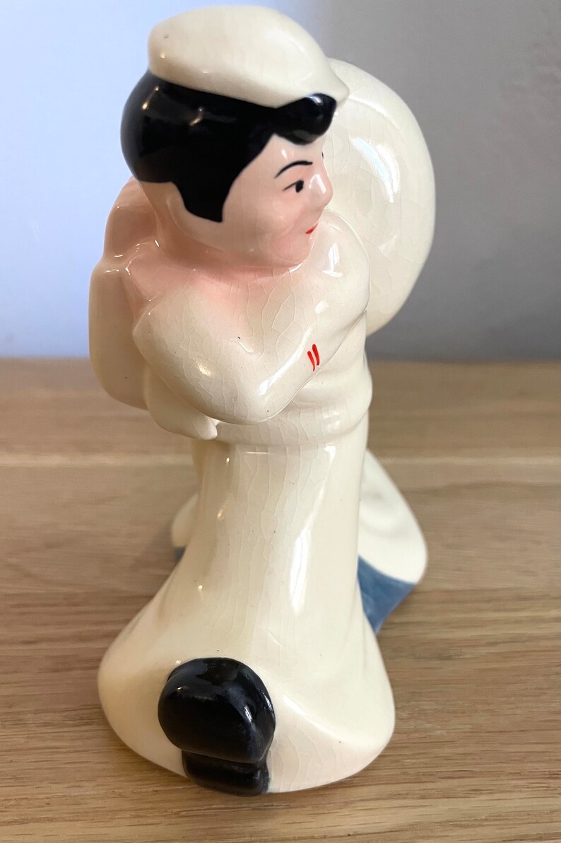 Op de afbeelding: Een keramiek beeldje van een matroos met een witte hoed, zwart haar en een wit uniform. De matroos houdt een groot wit voorwerp in zijn rechterhand. Het beeldje is geschilderd met rode accenten op het uniform.