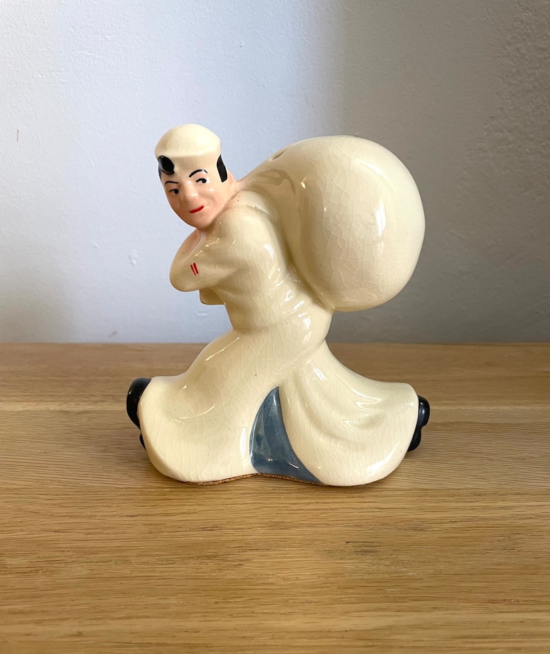 Op de afbeelding: Een keramiek beeldje van een matroos die een grote zak op zijn rug draagt. Het beeldje is geschilderd in tinten wit, blauw en zwart.
