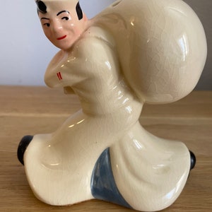 Op de afbeelding: Een keramiek beeldje van een matroos die een grote zak op zijn rug draagt. De matroos draagt een witte hoed en een wit shirt met een blauwe kraag. Het beeldje is geschilderd in tinten wit, blauw en zwart.