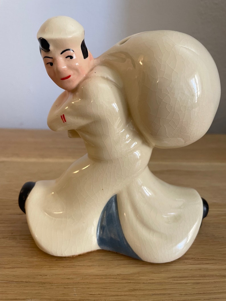 Op de afbeelding: Een keramiek beeldje van een matroos die een grote zak op zijn rug draagt. De matroos draagt een witte hoed en een wit shirt met een blauwe kraag. Het beeldje is geschilderd in tinten wit, blauw en zwart.
