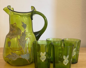 Puede incluir: Una jarra de vidrio verde y cuatro vasos a juego. La jarra tiene un diseño floral con detalles dorados. Los vasos presentan un diseño de conejito blanco. El conjunto se exhibe sobre una superficie de madera.