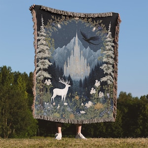 Könnte beinhalten: Gewebte Wandteppichdecke mit einer Fantasielandschaft mit einem weißen Schloss, einem Drachen und einem weißen Hirsch. Die Decke hat einen schwarzen Hintergrund und einen Fransenrand. Das Design umfasst Bäume, Blumen und Berge.