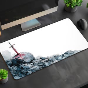 May include: A white rectangular mousepad featuring a watercolor illustration of a sword embedded in a rock. The sword has a red blade, and the rock is surrounded by blue and gray stones. The design is set against a white background.