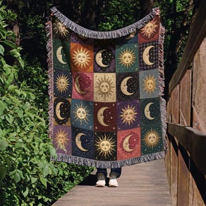 May include: A woven tapestry blanket featuring a repeating pattern of suns and moons in various colors. The blanket has a fringed edge and is held up against a natural background. The design includes celestial imagery.