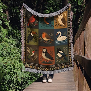 Peut inclure: Une couverture tissée avec un motif de grille d'illustrations d'oiseaux colorés, dont un cardinal et un toucan. La couverture a un fond sombre, une bordure décorative et des franges.
