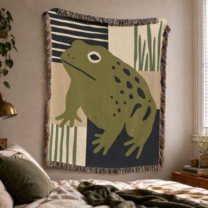 May include: A woven throw blanket featuring a large olive green frog with dark spots. The background is divided into geometric sections of cream, black, and olive green, including striped patterns and vertical lines. The edges have a fringed border.