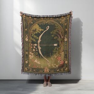 May include: A tapestry throw blanket with a forest scene and a bow and arrow design. The blanket has a dark green background with floral and tree details, and a fringed edge. The bow and arrow are white.