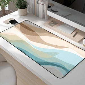 May include: A rectangular desk mat with a wavy design in shades of blue, beige, and brown. The mat has a black border and is placed on a white desk. A smartphone and other office supplies are visible.