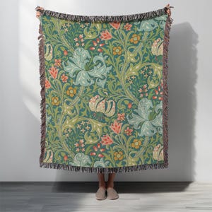 Puede incluir: Una manta tejida con un estampado floral en tonos turquesa, verde, melocotón y crema. El diseño presenta flores y follaje estilizados. La manta tiene un borde con flecos.