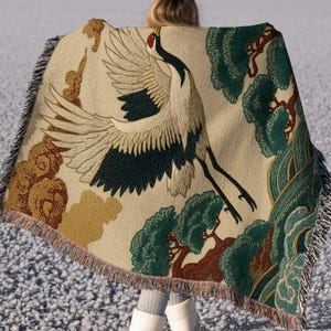 May include: A woven throw blanket featuring a large red-crowned crane in flight, surrounded by stylised green pine trees and golden-brown clouds on a cream-coloured background. The blanket has a textured weave and fringed edges on the sides.